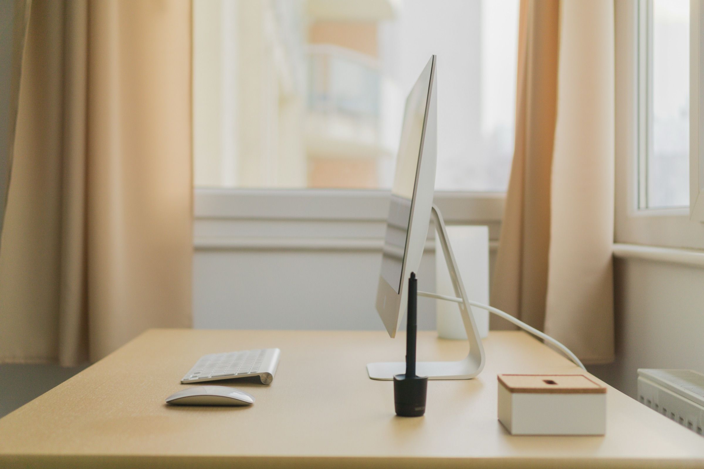 A tidy desk with a computer, keyboard and pen pot beside a bright window