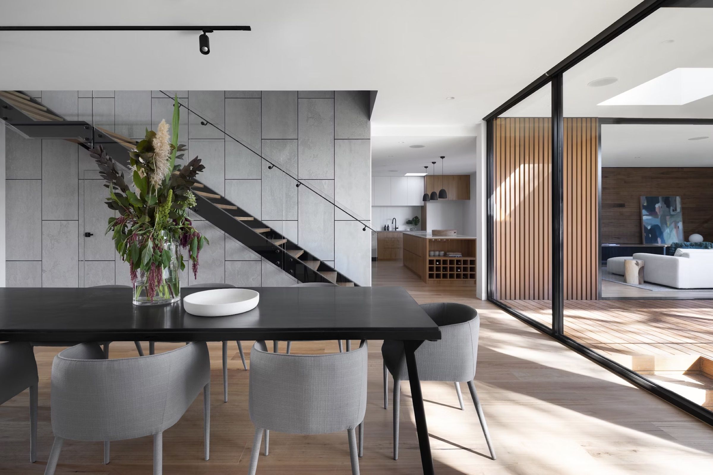 Dining area with a long table beside a floating staircase and a feature wall